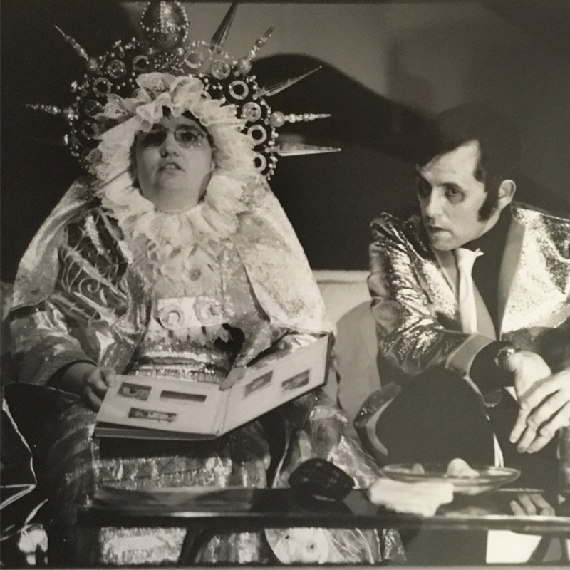 2 actors from Lung Ha Theatre Company are sat on a couch looking at a photo album. 1 is costumed as Mary wearing an elaborate headdress and the other is Elvis Presley in a shiny jacket. Costumes designed by Alison Brown.