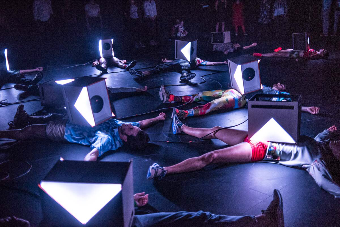 8 dancers from Scottish Dance Theatre are lying on the floor with speakers on their chests. They are watched by an audience as they perform contemporary dance show Looping wearing costumes made from Timorous Beasties fabric and designed by Alison Brown Costume Designer.