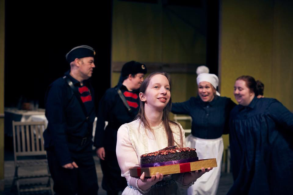 5 actors from Lung Ha Theatre Company on stage at the Traverse Theatre performing Three Sisters by Anton Chekhov. 2 of the sisters are pictured with 1 servant and 2 soldiers all wearing costumes designed by Alison Brown Costume Designer. The  actress in the foreground is carrying a cake.