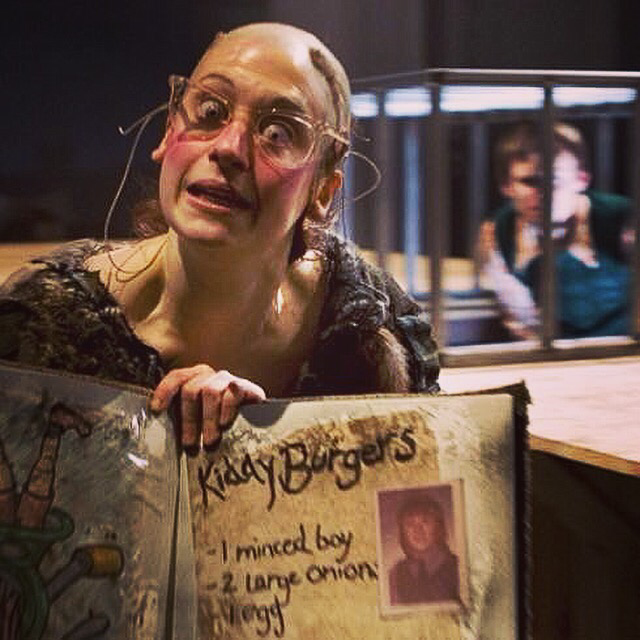 The Witch from Hansel & Gretal is wearing classes and has a bald head. She is holding up a cookery book with a recipe for kiddy burgers. Behind her can be seen Hansel in a cage.