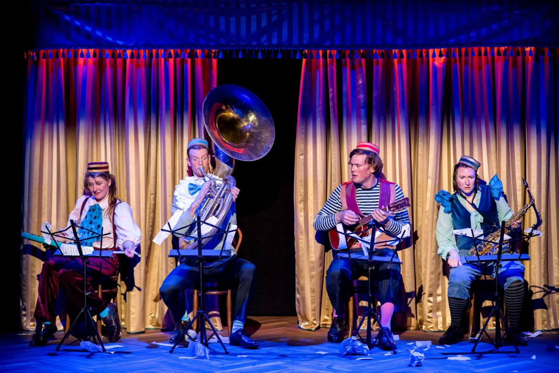 4 performers are sitting looking confused with various musical instruments and music stands performing Whirlygig in costumes designed by Alison Brown Costume Designer. They are sitting in front of a gold curtain with tassles.